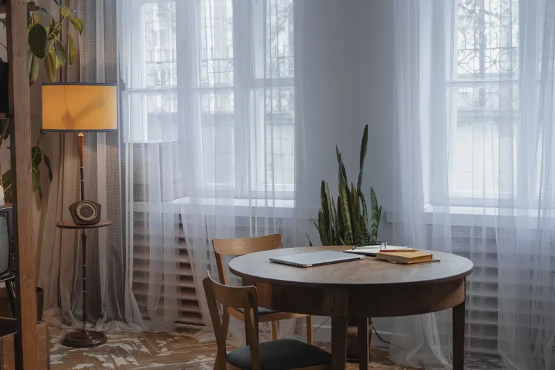 Mesa de madera junto a los ventanales, con visillos